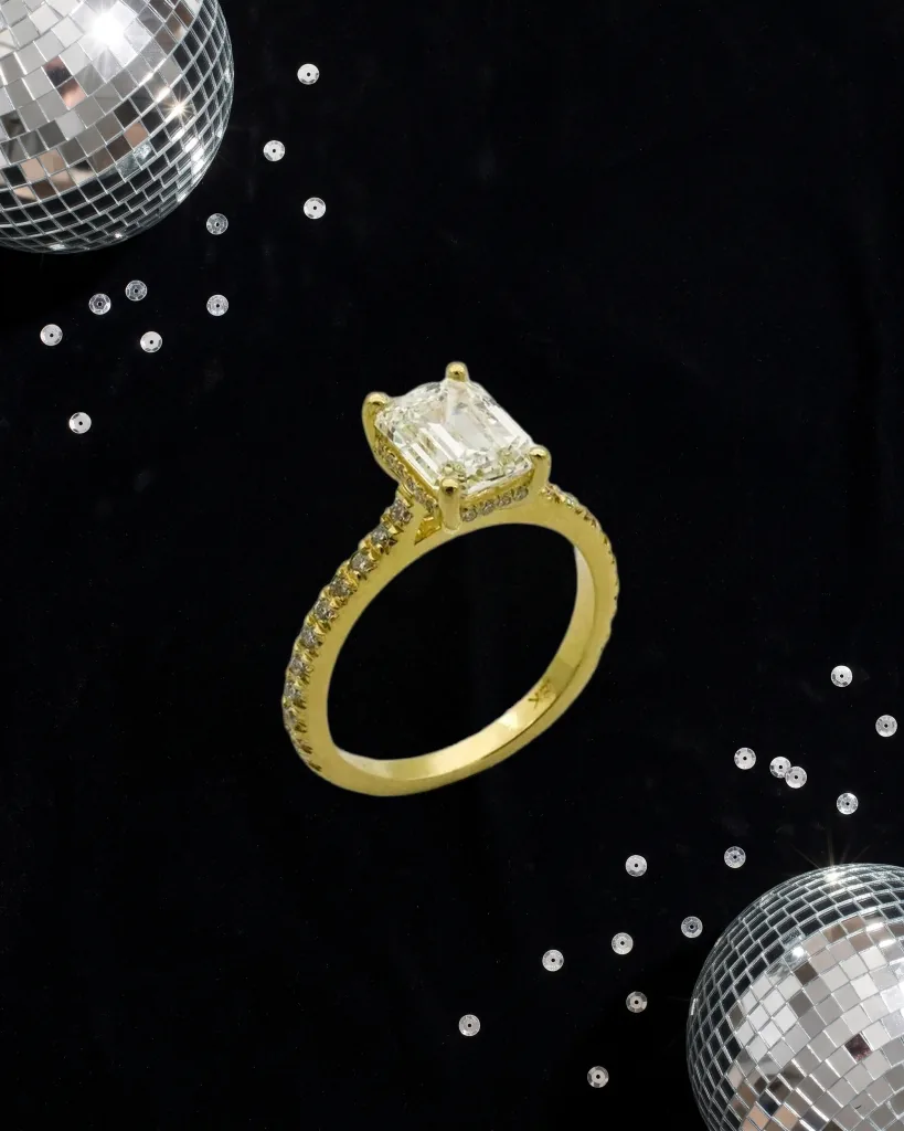 Gold ring with a square stone on black velvet between two cropped disco balls and silver sequins