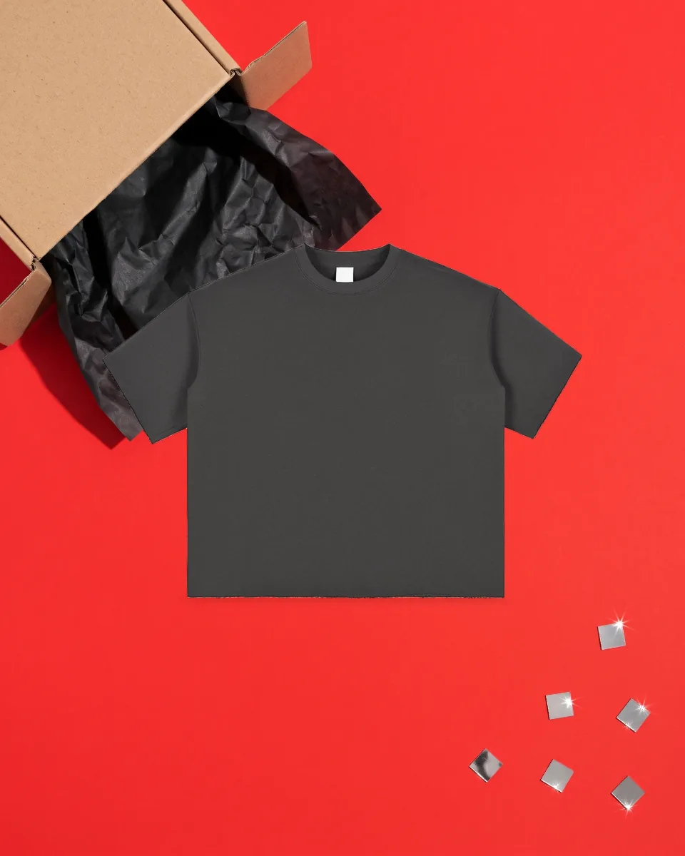 Dark grey T-shirt laid flat on red paper beside an open kraft box with black tissue, seen from above