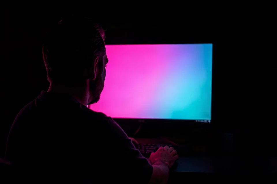 Person at a desk facing a monitor that glows pink and cyan in a dark room