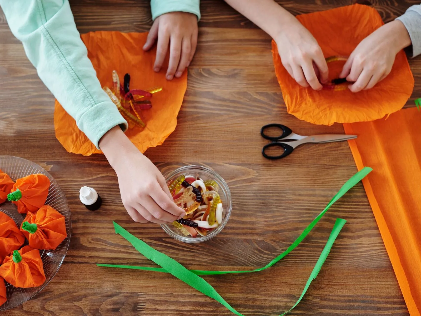 Children's hands wrapping gummy worms in orange tissue paper to make pumpkin treat bundles