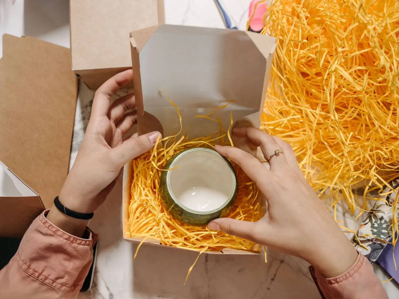 Hands placing a ceramic mug in orange paper shred inside a small shipping box