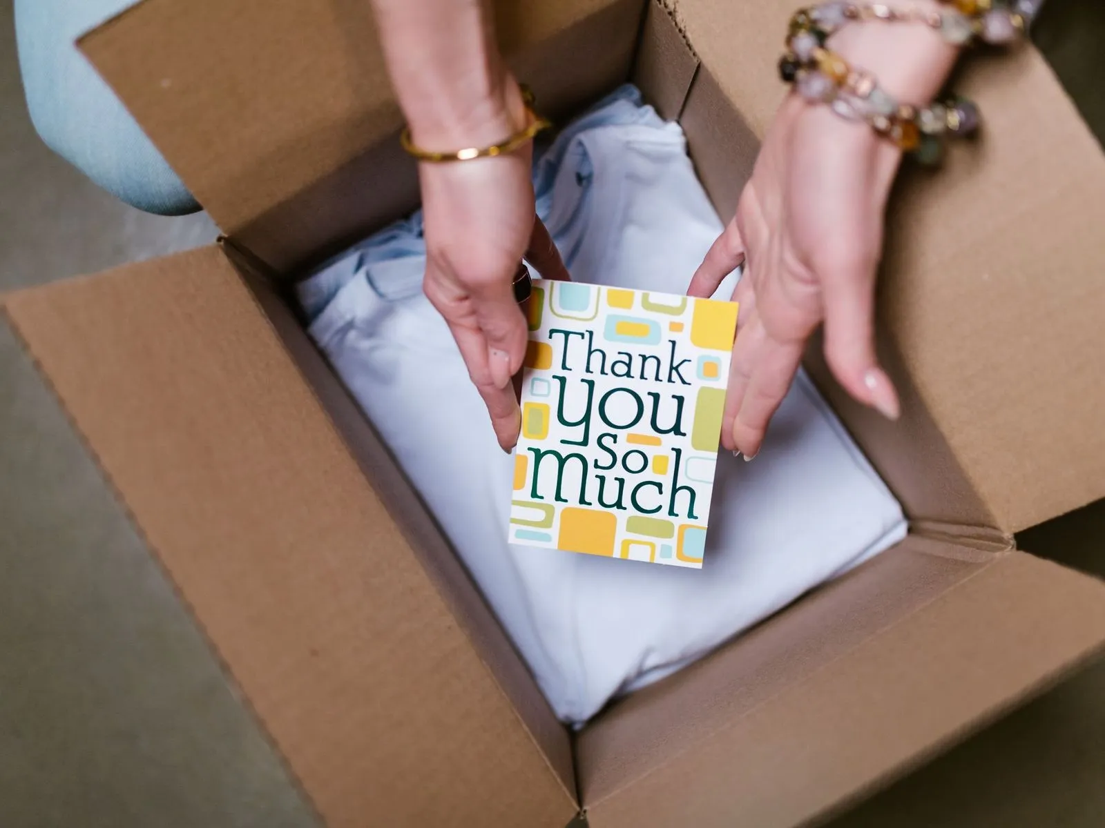 Hands holding a Thank You So Much card above a folded shirt in a shipping box