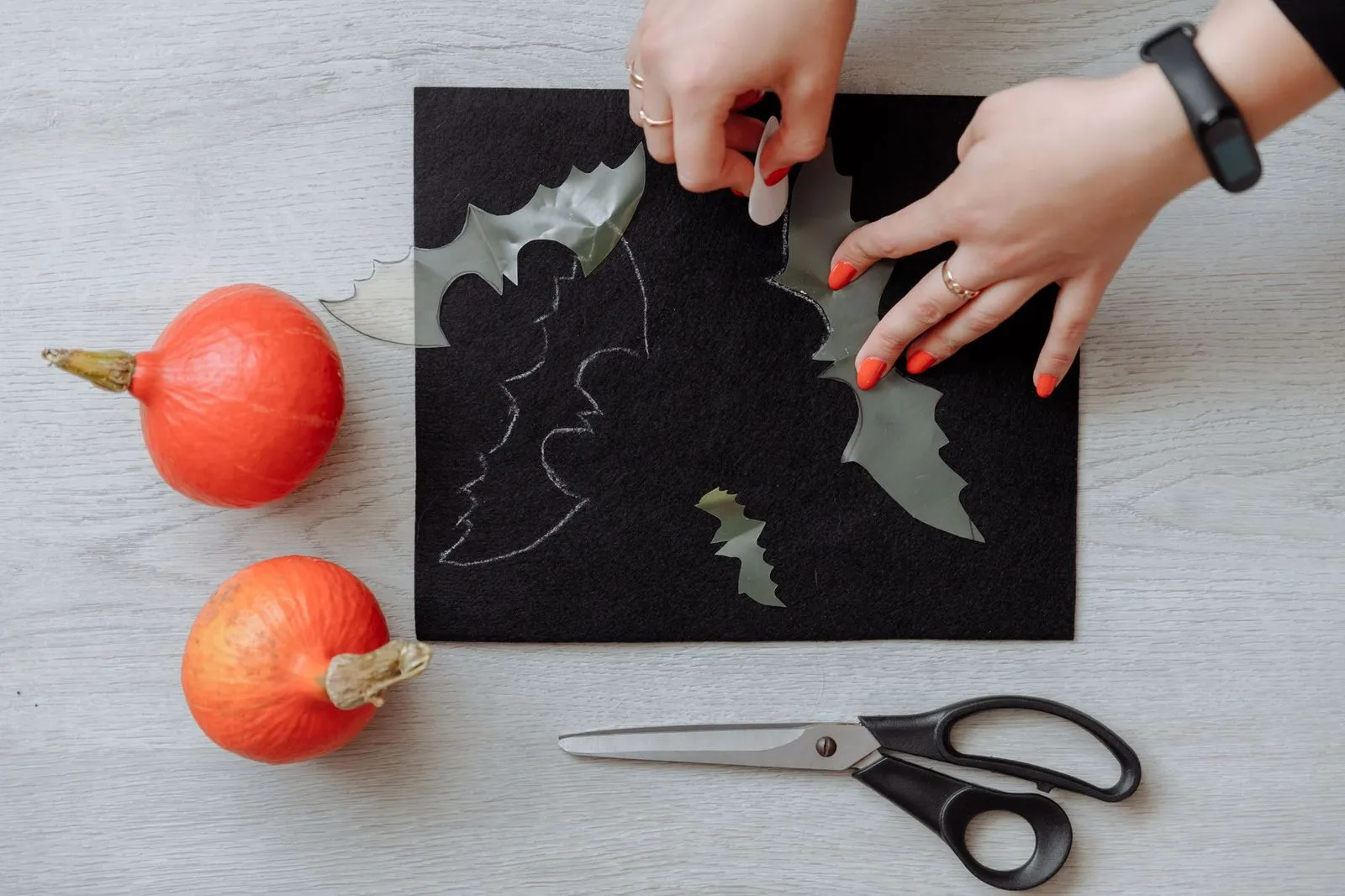 Hands cutting black felt bat shapes beside two small orange pumpkins and scissors