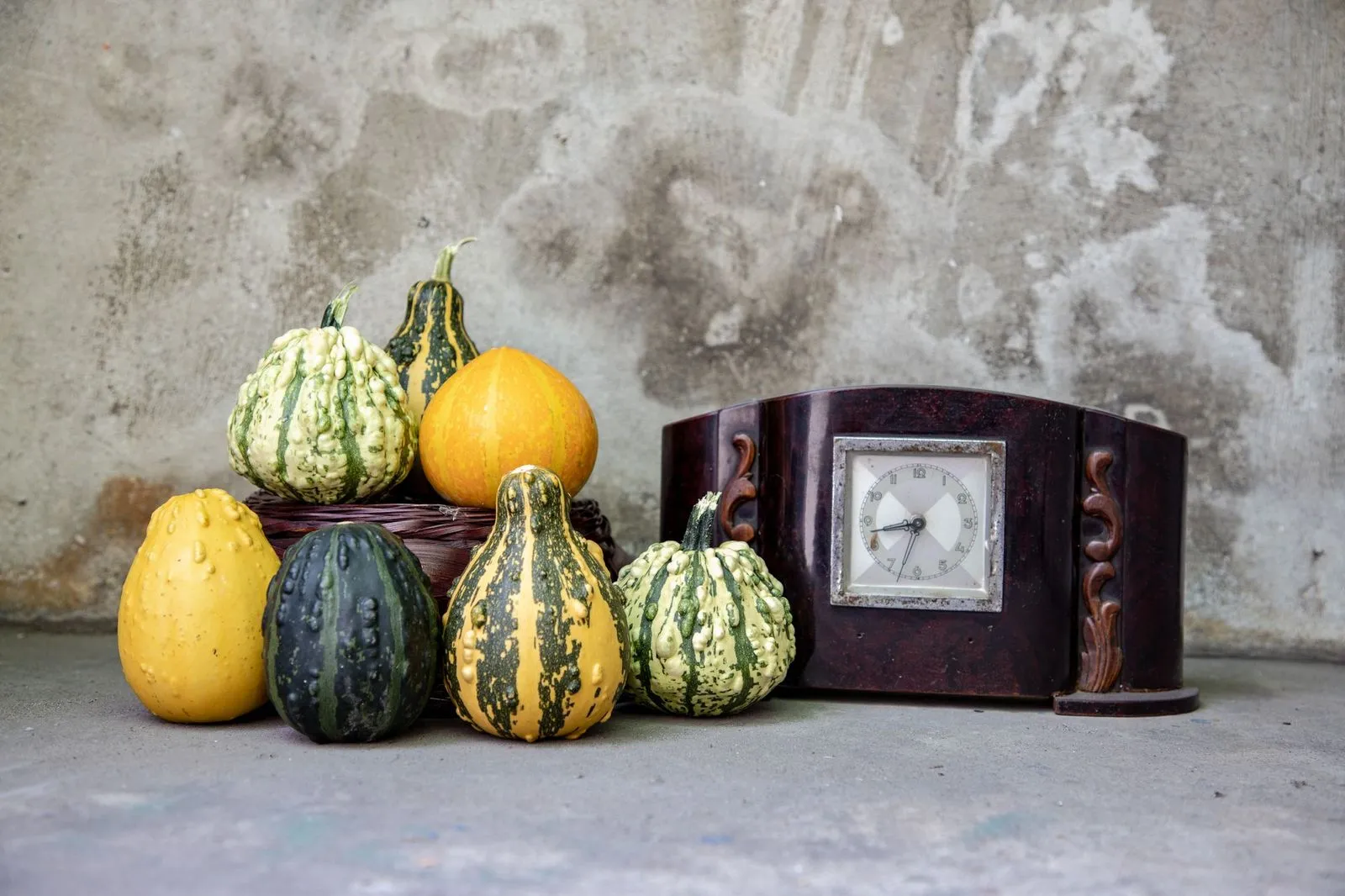 Striped gourds and yellow squash beside a vintage wooden mantel clock on concrete