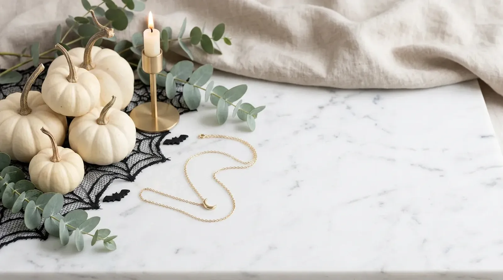 Gold crescent-moon necklace on white marble with white pumpkins and spiderweb lace, space on the right